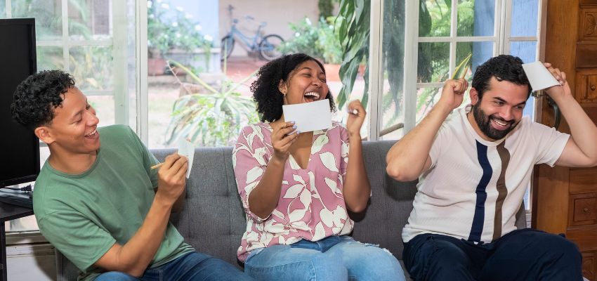 Three friends laughing and playing a card game on the couch as part of fun bible games for adults