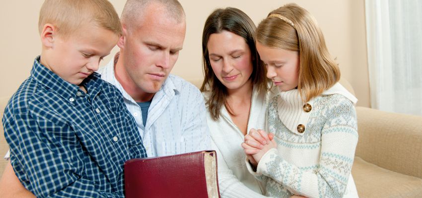 Parents and children gathered in living room sharing night prayers for family with the Bible.