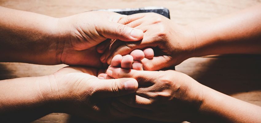 Diverse hands of different ages and skin tones stacked together in a circle of unity over an open Bible – representing community support and collective fertility prayers lifted up in faith for those hoping to conceive or carry a child.