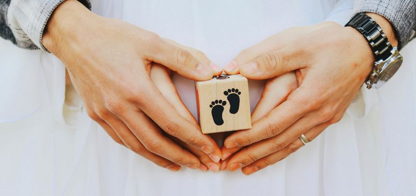 Couple's hands forming a heart shape around a small wooden block engraved with baby footprints, symbolizing longing and hope – illustrating fertility prayers for the gift of new life, pregnancy, and the miracle of parenthood.
