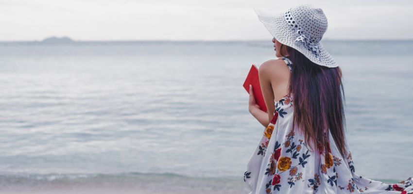 Woman by the shore holding a book, finding hope in bible verses about waiting for love.