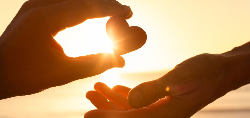 Hands holding a heart symbol against the sunlight, representing love, compassion, and kindness inspired by Bible verses.