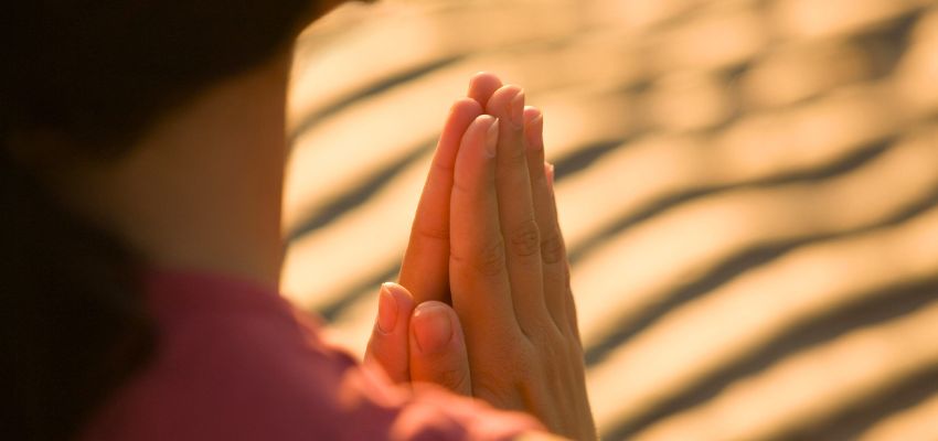 Woman with hands clasped together on sandy beach at sunrise, embodying the peace and power found in a bible verse for strength during personal reflection.