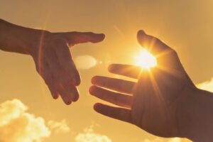 Silhouette of two outstretched hands nearly touching against a glowing sunset sky with a wooden cross in the background, symbolizing compassion and connection inspired by bible verses about helping the poor.