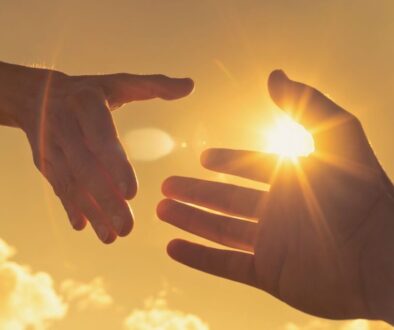 Silhouette of two outstretched hands nearly touching against a glowing sunset sky with a wooden cross in the background, symbolizing compassion and connection inspired by bible verses about helping the poor.