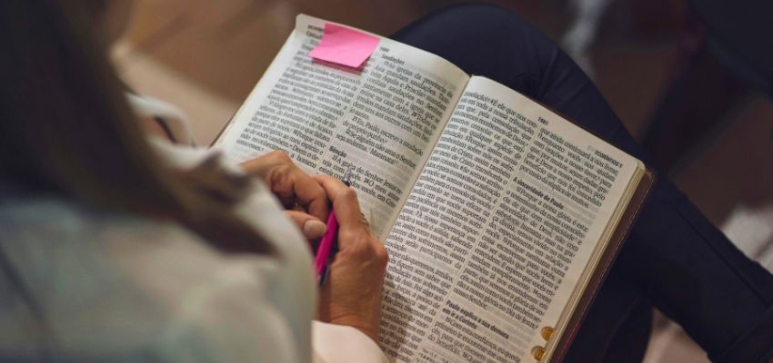 Close-up of hands holding an open Bible with pink sticky notes and underlining on scripture pages, representing personal study of bible verses about helping the poor and living out generosity.
