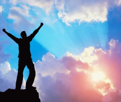 Powerful silhouette of a man standing atop a mountain peak with arms raised high in victory and praise, backlit by radiant blue and orange sunset clouds, evoking triumph, resilience, and divine empowerment found in bible verses about love and strength.