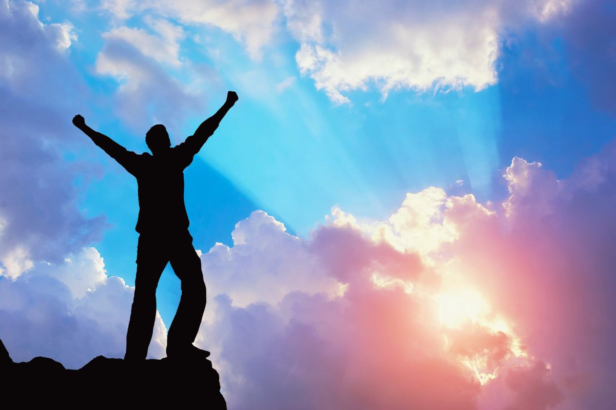 Powerful silhouette of a man standing atop a mountain peak with arms raised high in victory and praise, backlit by radiant blue and orange sunset clouds, evoking triumph, resilience, and divine empowerment found in bible verses about love and strength.