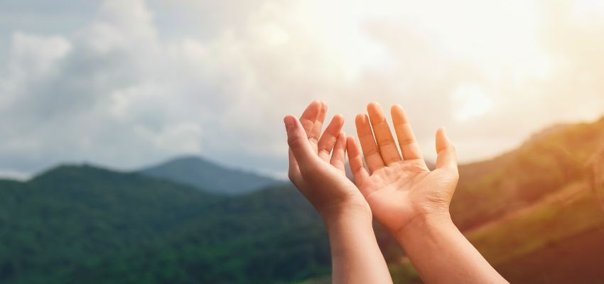 Pair of open hands raised palms-up toward a glowing sunrise over lush mountain ridges and valleys, bathed in warm golden light, symbolizing surrender, hope, and renewal often expressed in bible verses about love and strength.