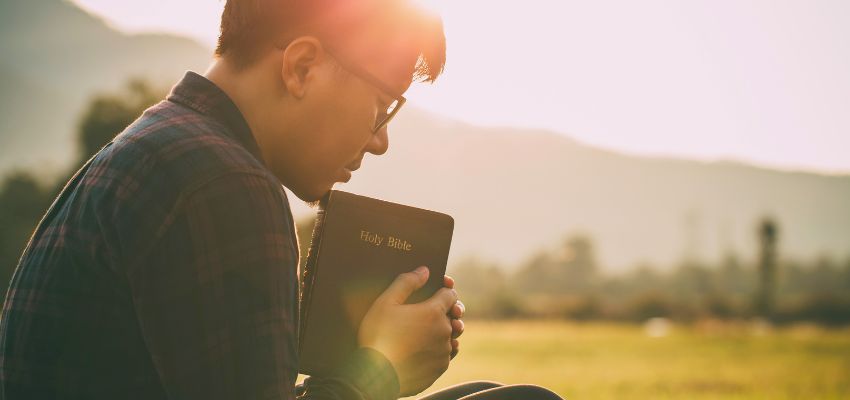 Thoughtful young man in a plaid shirt sitting on grass with mountains in the background, eyes closed in prayer while gently holding an open Holy Bible in his hands, representing personal reflection and comfort drawn from bible verses about love and strength.