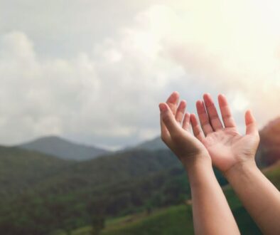 Open hands raised toward the sky in worship, person's palms facing upward against a scenic backdrop of lush green mountains and cloudy sunset sky, representing offering, trust, and responsible care of creation inspired by bible verses on stewardship.