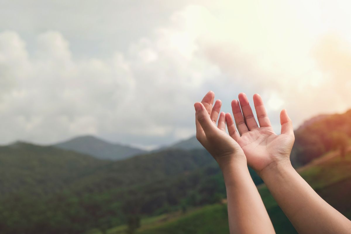 Open hands raised toward the sky in worship, person's palms facing upward against a scenic backdrop of lush green mountains and cloudy sunset sky, representing offering, trust, and responsible care of creation inspired by bible verses on stewardship.