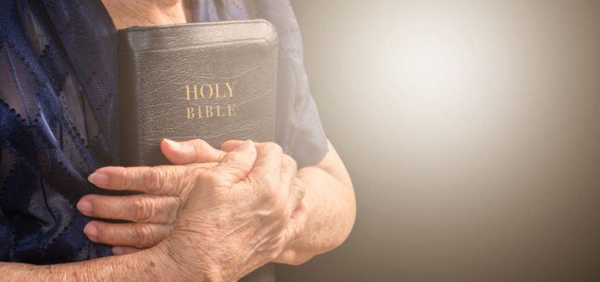 Elderly woman gently cradling a Holy Bible to her chest, close-up of wrinkled hands and navy blue lace-trimmed blouse, soft glowing light highlighting the leather-bound book titled HOLY BIBLE, evoking faithful stewardship of God's Word and life lessons from bible verses on stewardship.
