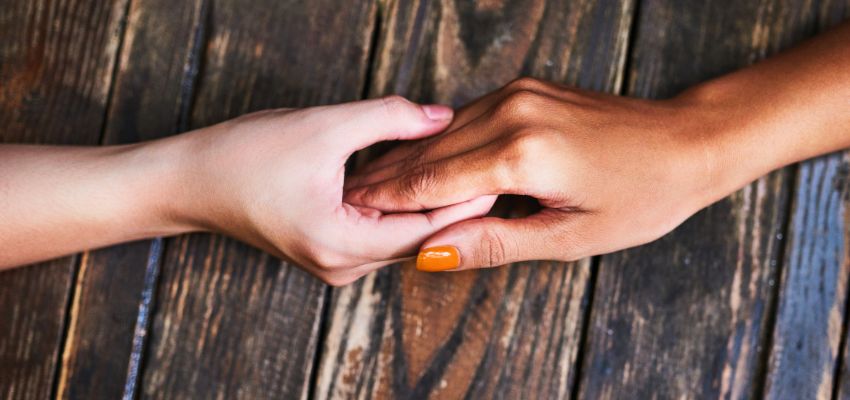 Close-up of two hands clasped together—one lighter-skinned and one darker-skinned—resting on a wooden table, symbolizing unity, support, and love as expressed in friendship scripture and biblical teachings on companionship