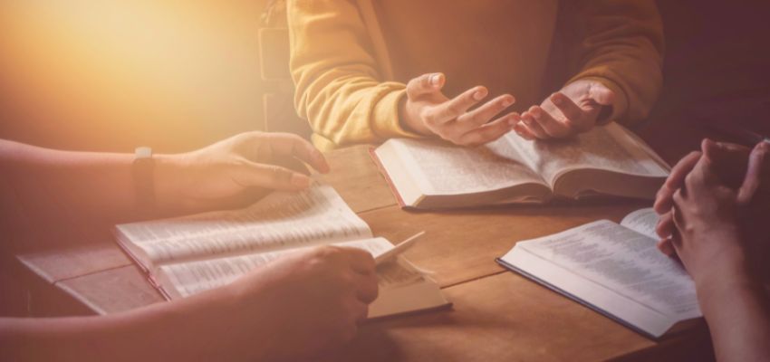 Warmly lit scene of a small group studying open Bibles together at a wooden table, hands gesturing in discussion and pointing to passages, evoking the spirit of friendship scripture shared in community and fellowship
