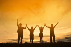 Joyful silhouette of four children holding hands and raising their arms in celebration against a glowing orange sunset sky, capturing the innocence, unity, and rejoicing found in friendship scripture and verses celebrating faithful companions