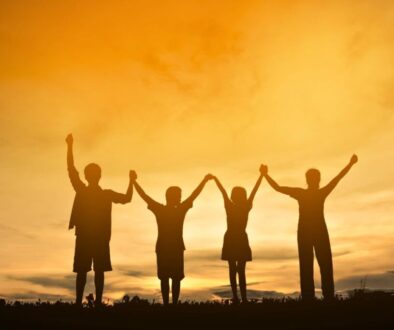 Joyful silhouette of four children holding hands and raising their arms in celebration against a glowing orange sunset sky, capturing the innocence, unity, and rejoicing found in friendship scripture and verses celebrating faithful companions