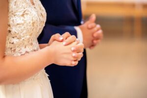Prayer for husband and wife: Close-up of a bride in a white lace wedding dress and groom in a suit holding hands together in prayer