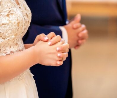 Prayer for husband and wife: Close-up of a bride in a white lace wedding dress and groom in a suit holding hands together in prayer