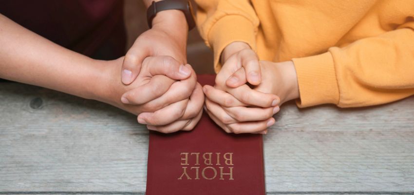 Prayer for husband and wife: Two pairs of hands clasped together in prayer resting on a Holy Bible, representing family faith and spiritual connection