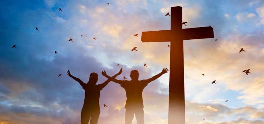 Silhouette of a couple praising with arms raised before a large cross at sunset, evoking hope and inspiration for a new year prayer for family.