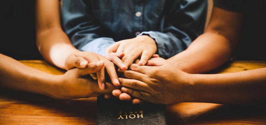 Close-up of adult and children’s hands joined in unity during prayers for children over a Holy Bible.