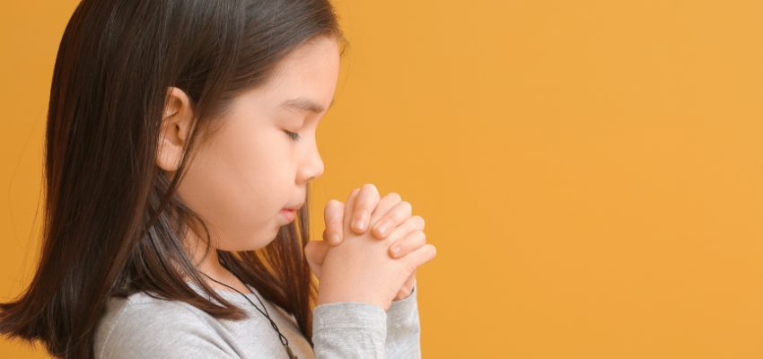 Young girl with eyes closed and hands clasped in quiet prayers for children against a warm background.